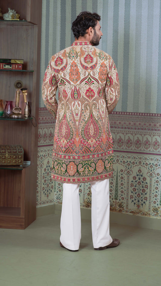 A man viewed from behind wearing a beige kurta with intricate multicolor paisley embroidery in red, green, and gold, paired with white trousers, standing in a decorated room with patterned wallpaper.