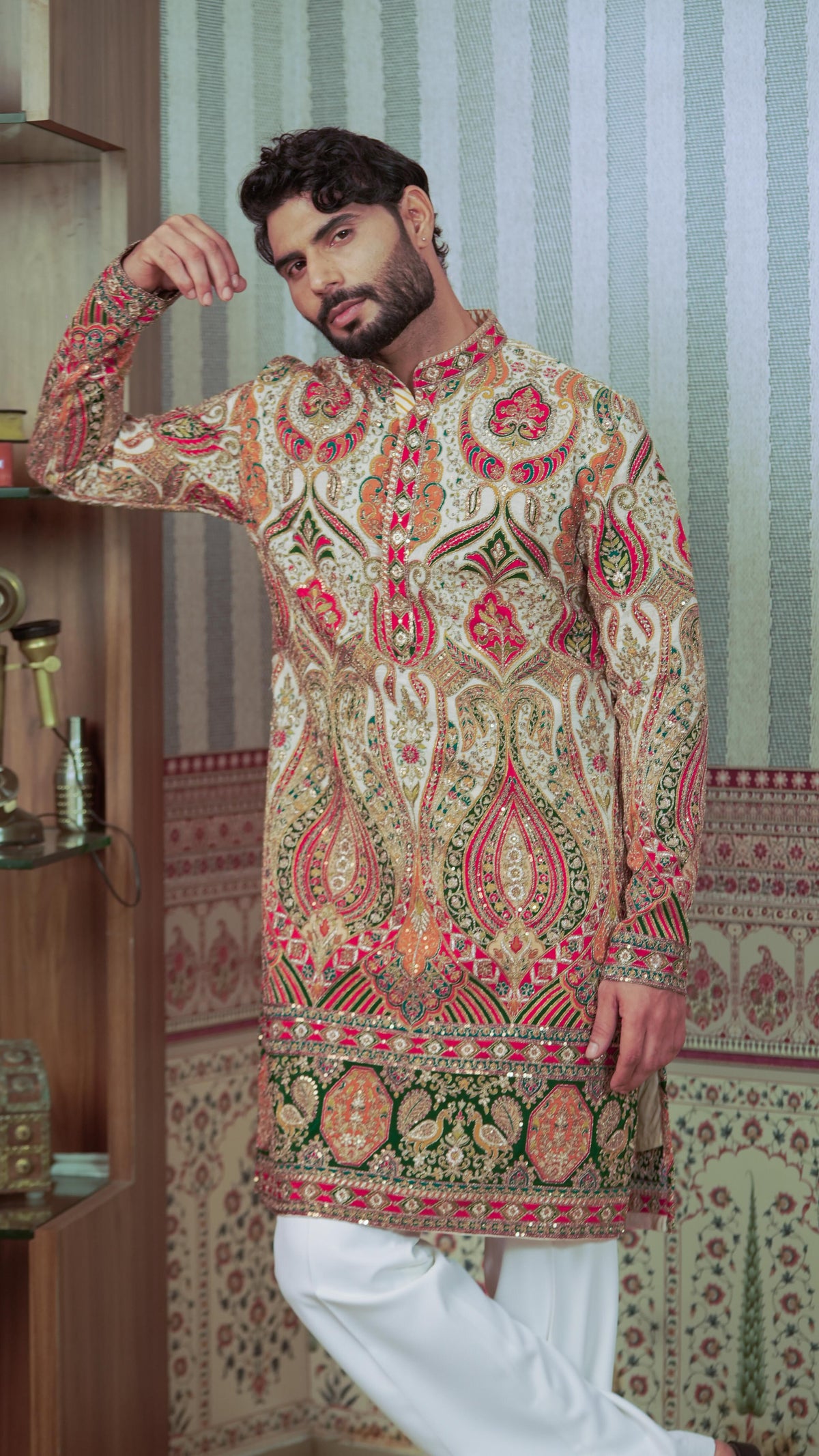 A man wearing a long-sleeved, mandarin-collar kurta with an intricate multicolor paisley and floral pattern in beige, red, green, and gold, paired with white trousers, standing in front of a patterned wallpaper background.