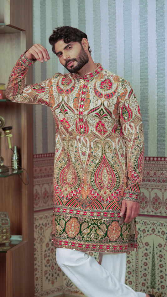 A man wearing a long-sleeved, mandarin-collar kurta with an intricate multicolor paisley and floral pattern in beige, red, green, and gold, paired with white trousers, standing in front of a patterned wallpaper background.