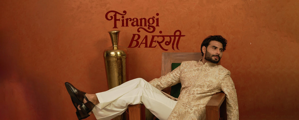 A man in a beige patterned sherwani and white trousers reclines on a wooden chair against a terracotta wall, with the text 'Firangi BAeRangi' written above and a gold vase beside him.