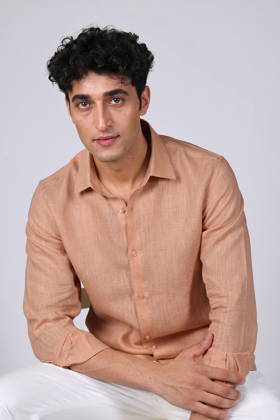 Peach Sandstone pure linen shirt for men, seated portrait with arms crossed, classic spread collar and buttoned cuffs showing linen texture, styled with white trousers, by BWO
