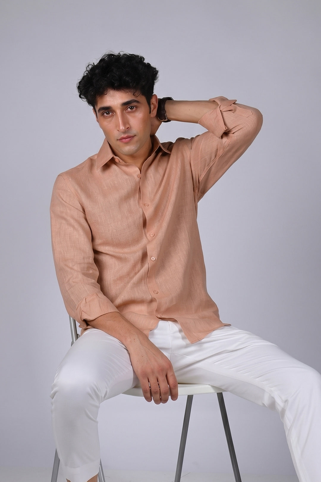 Peach Sandstone pure linen shirt for men, seated pose with hand behind neck, classic spread collar and button placket, styled with white trousers, by BWO