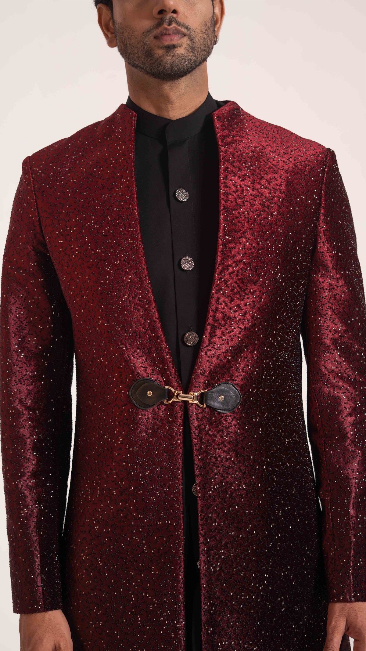 A man wearing a sparkling deep red sherwani jacket over a black kurta, featuring a decorative gold-toned tie closure at the waist.