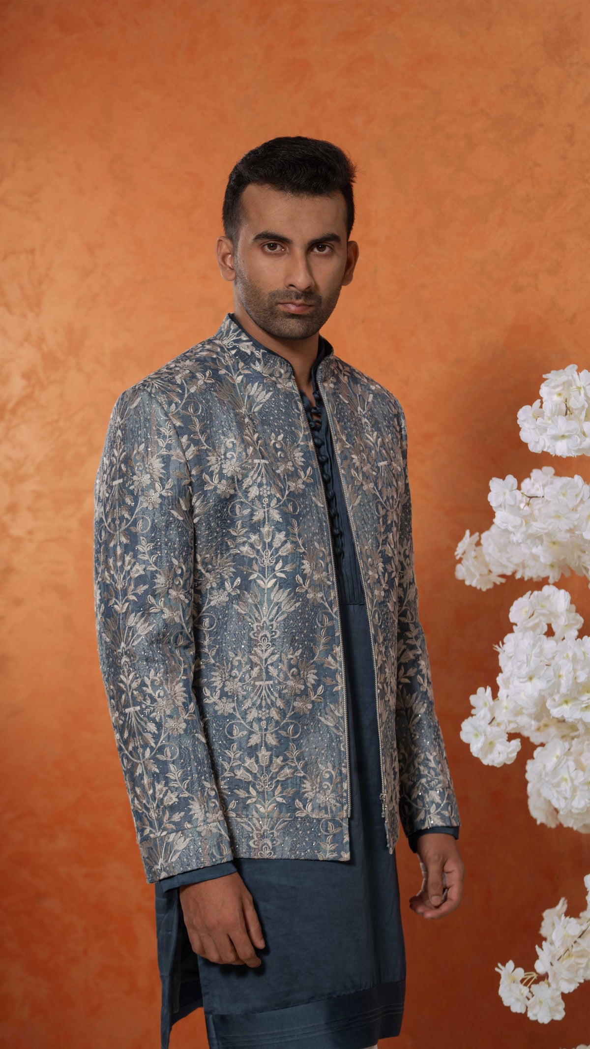 A man wearing a blue and beige patterned sherwani jacket over a dark blue kurta, standing against a textured orange background with white decorative flowers on the right.