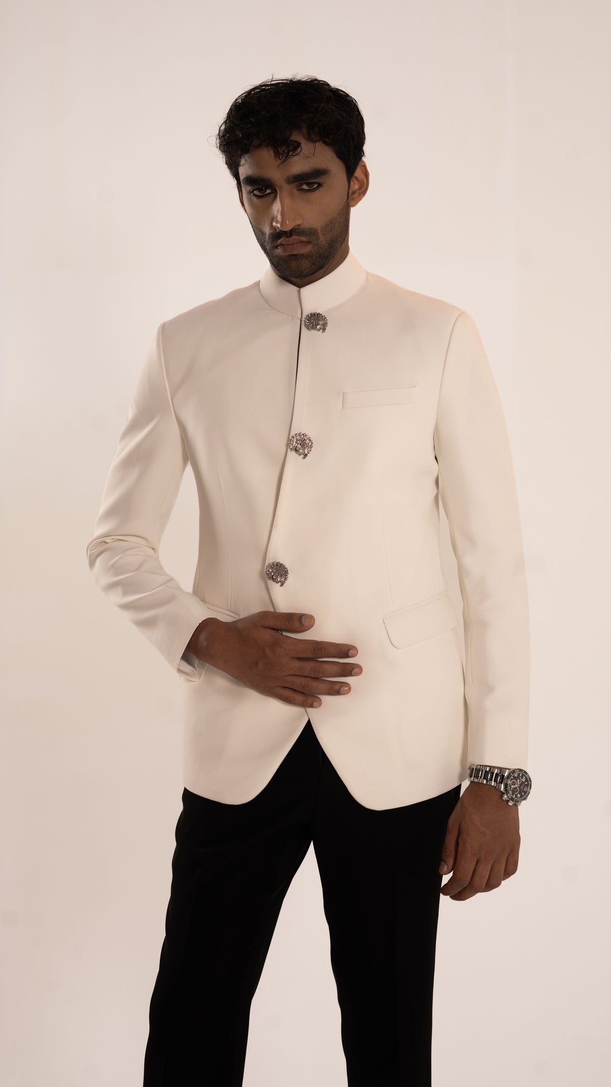 A man modeling a cream-colored traditional Indian jacket (bandhgala) with silver decorative buttons, paired with black trousers, against a plain off-white background.
