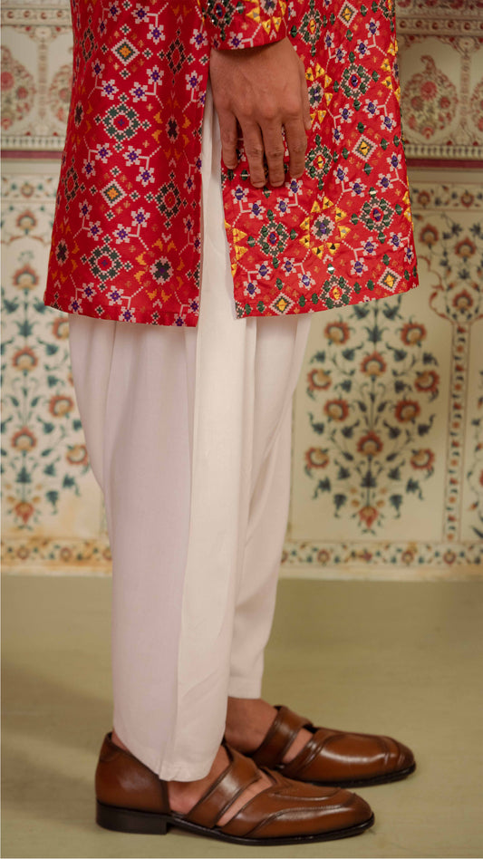 A person wearing a bright red patterned kurta with white pants and brown leather loafers, standing indoors against a patterned wall.