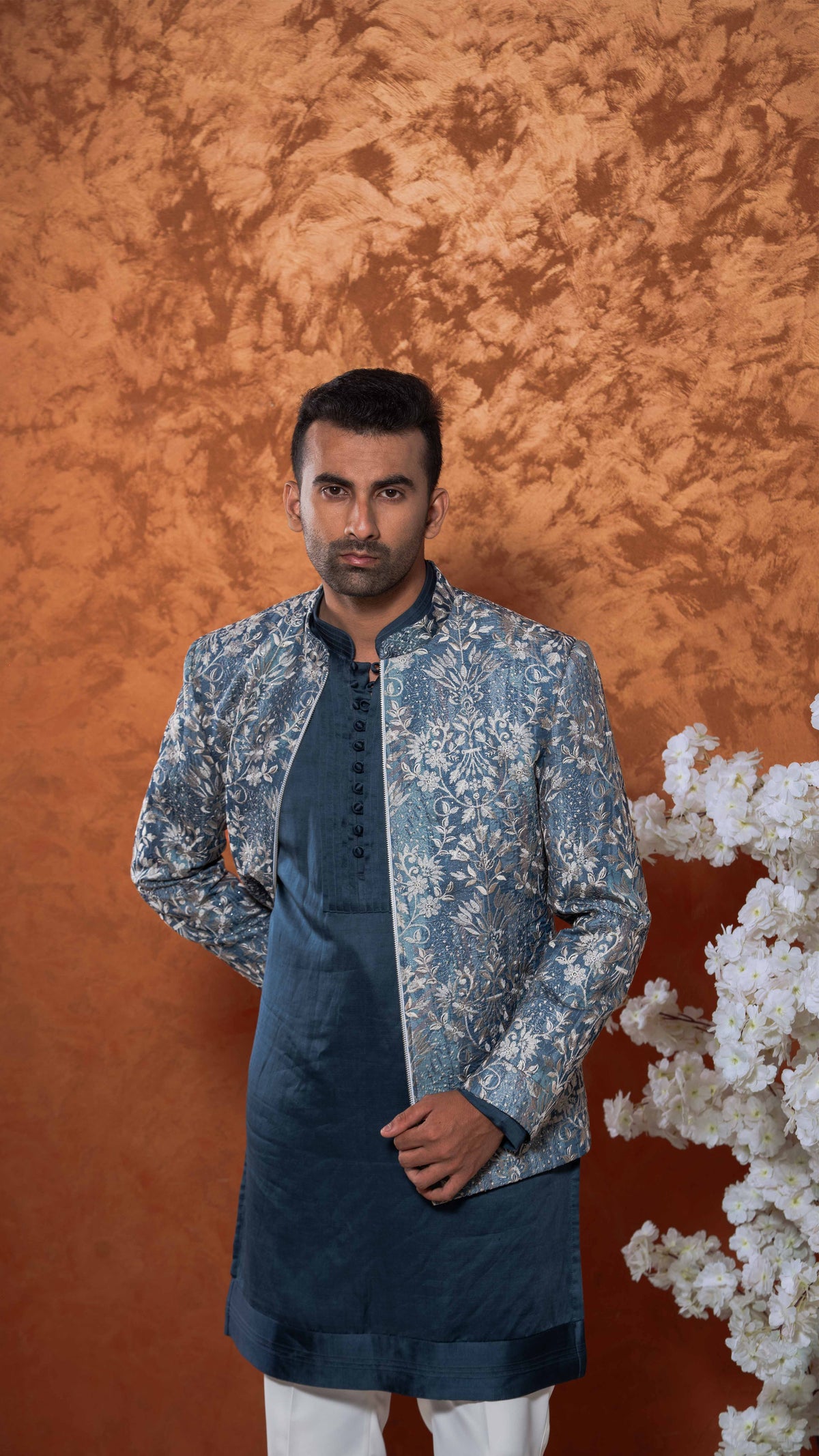 A man modeling a traditional Indian outfit consisting of a navy blue kurta with a light blue and white floral patterned sherwani jacket, paired with white trousers, set against a textured orange-brown backdrop with white decorative flowers on the right.