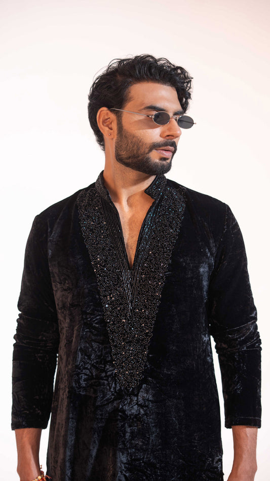 A man wearing a black velvet kurta with a heavily embellished, beaded black shawl collar and dark sunglasses, photographed against a clean white studio background.