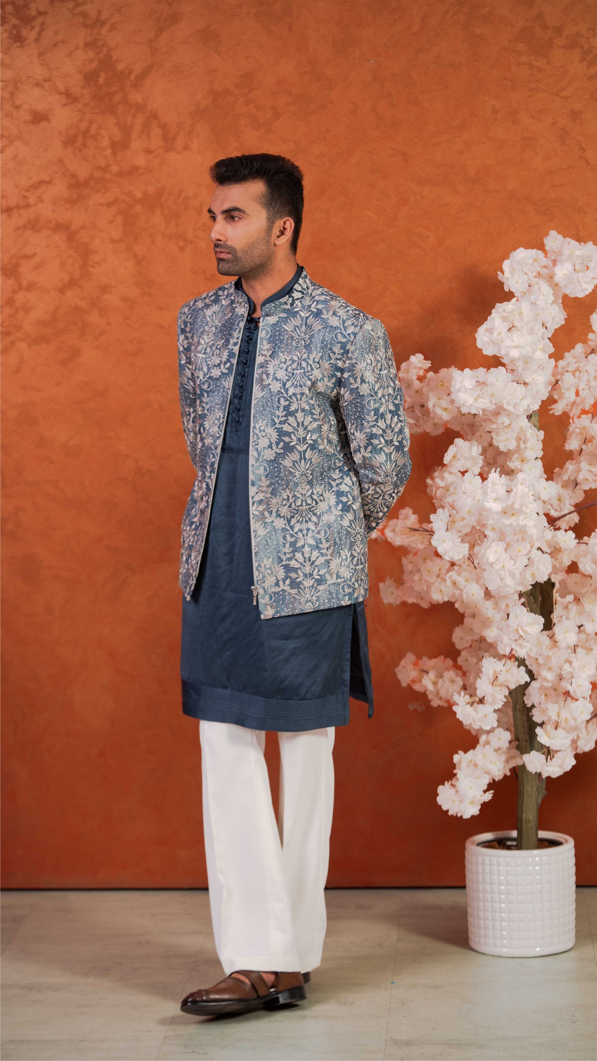 A man modeling a traditional Indian outfit consisting of a navy blue kurta, white trousers, and a blue and white patterned jacket, standing against a textured orange wall next to a potted flowering tree.