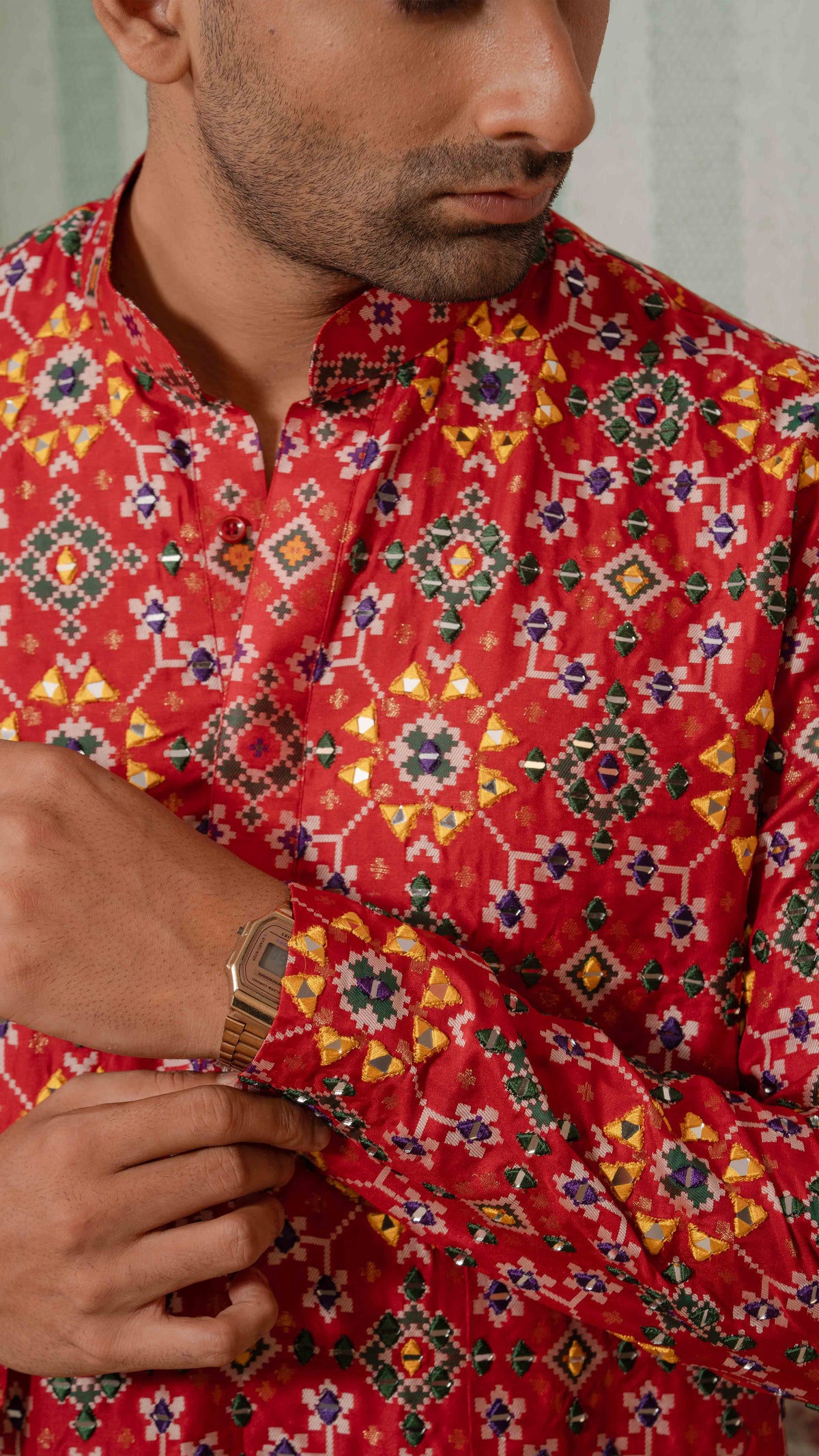 A close-up of a man wearing a red long-sleeve kurta with a colorful geometric and floral pattern, adjusting the cuff of the sleeve.