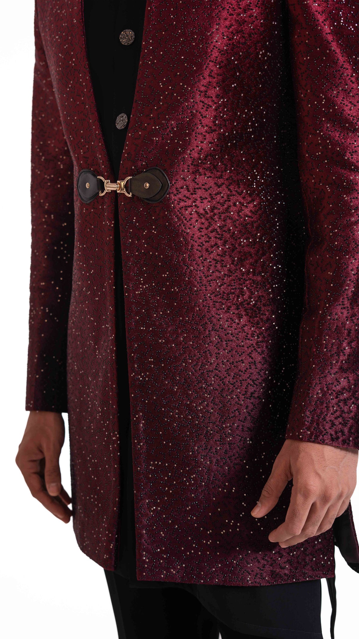 A close-up of a person wearing a sparkling burgundy jacket with a black shirt underneath and black trousers. The jacket features a unique toggle closure with two dark brown circular pieces connected by a gold-colored chain.