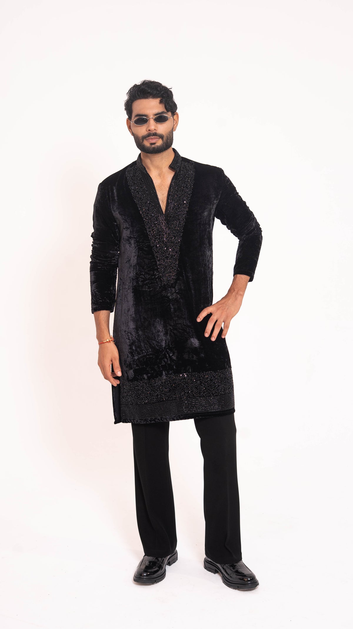 A man modeling a black velvet kurta jacket with sequined trim, paired with black trousers and black shoes, against a plain white studio background.