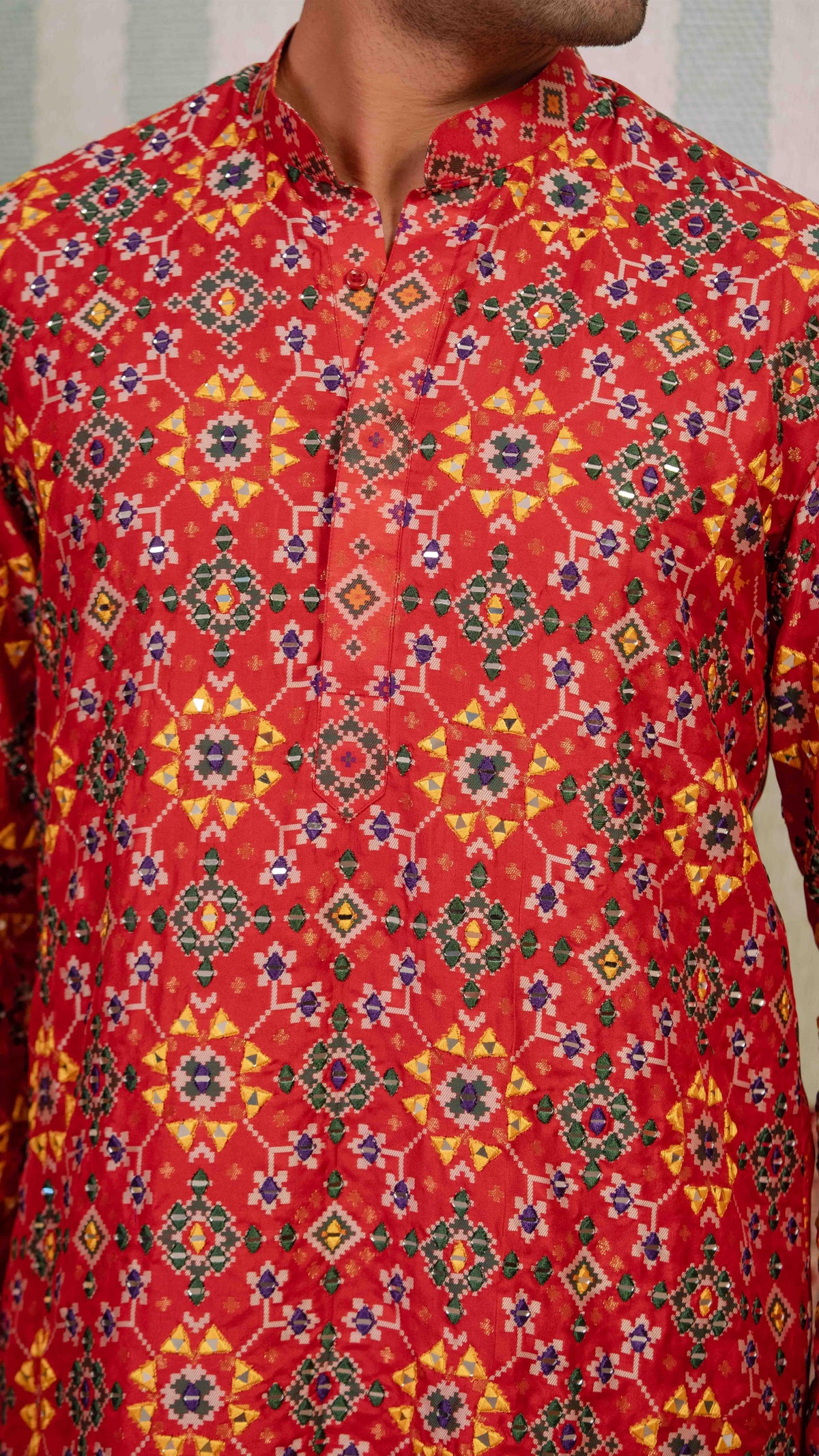 A close-up of a man wearing a red patola kurta with a detailed geometric and floral pattern in yellow, green, and blue.