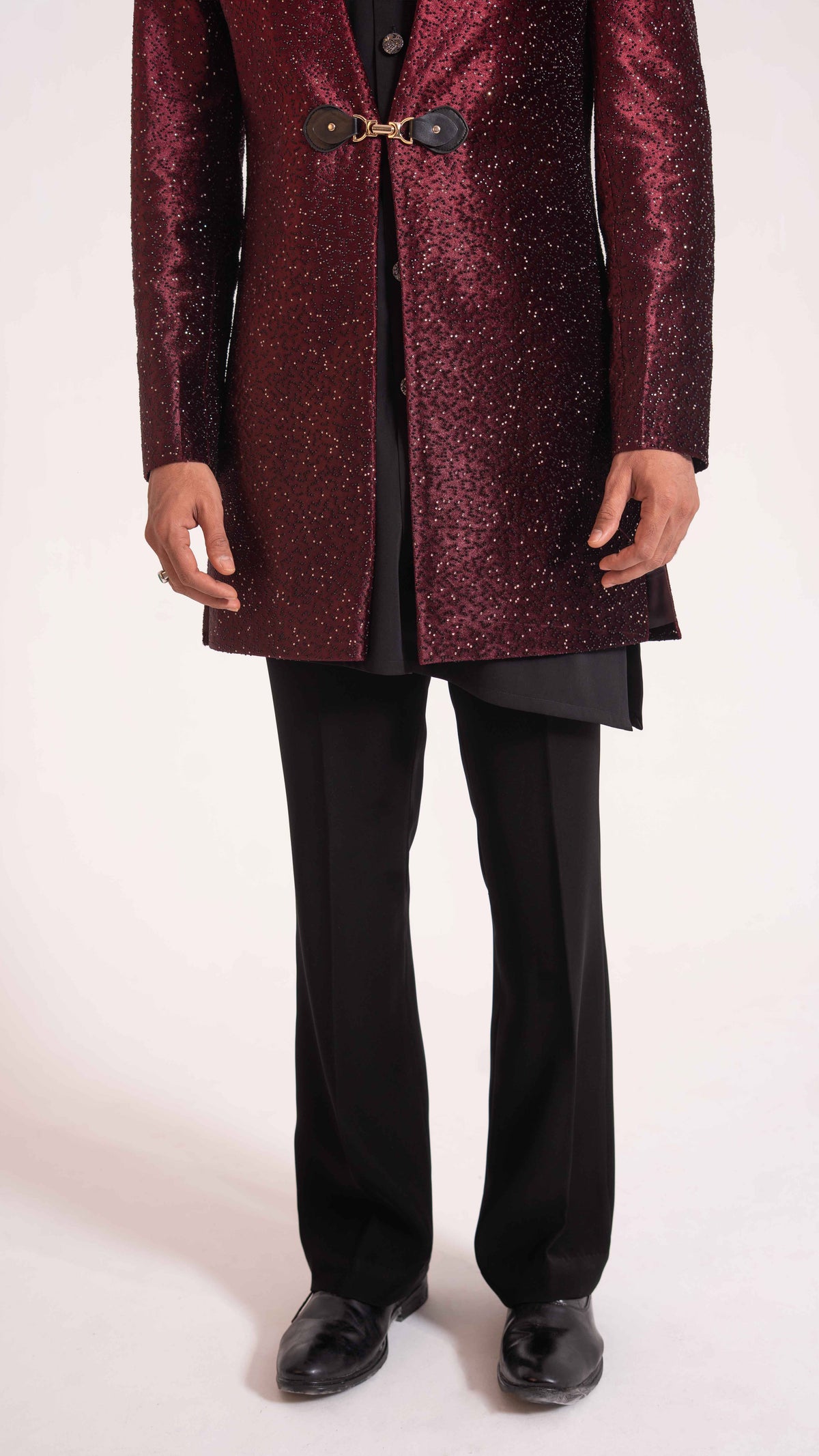 A man wearing a sparkling deep burgundy sequined long coat with a decorative gold-toned front detail, paired with black trousers and black shoes, photographed against a plain light background.