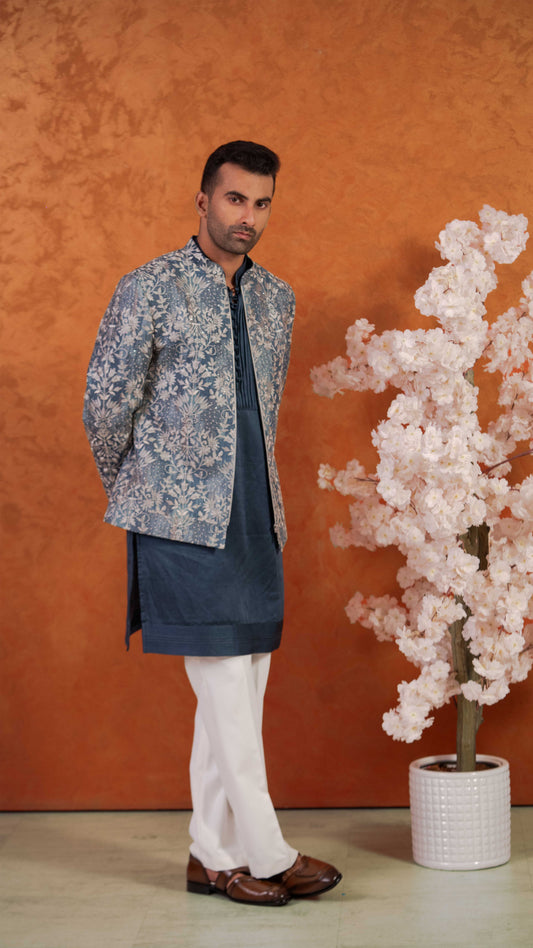 A man modeling a traditional Indian outfit consisting of a blue and white patterned jacket, a navy blue kurta, white trousers, and brown leather shoes, standing against a textured orange wall next to a decorative pink flowering tree.