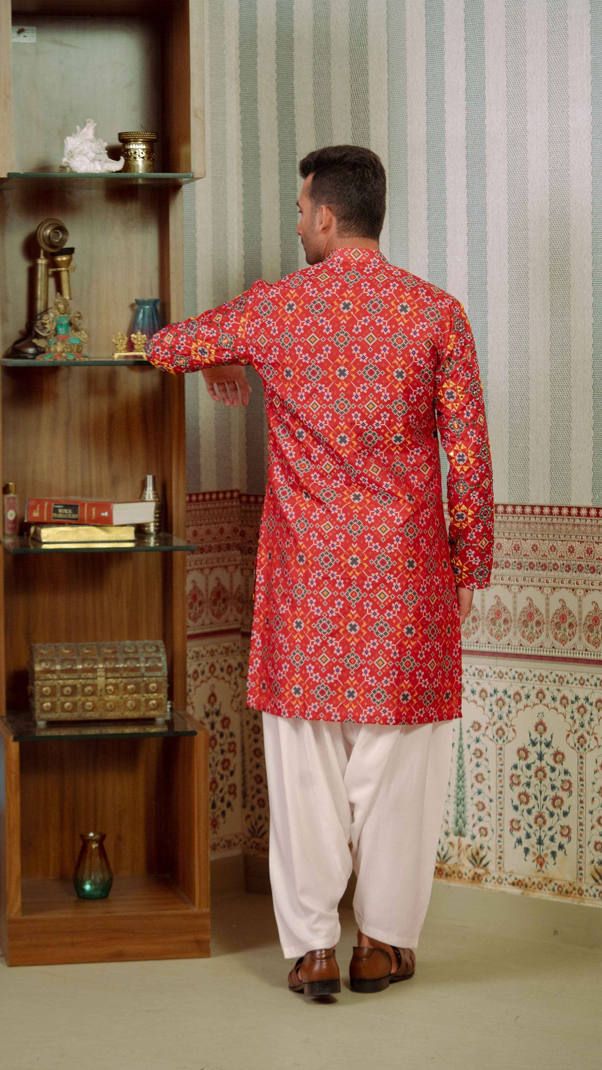 A man viewed from behind wearing a red long-sleeve kurta with a dense yellow, orange, and blue floral pattern, paired with white pants and brown shoes, standing in front of a wooden shelf and patterned wallpaper.