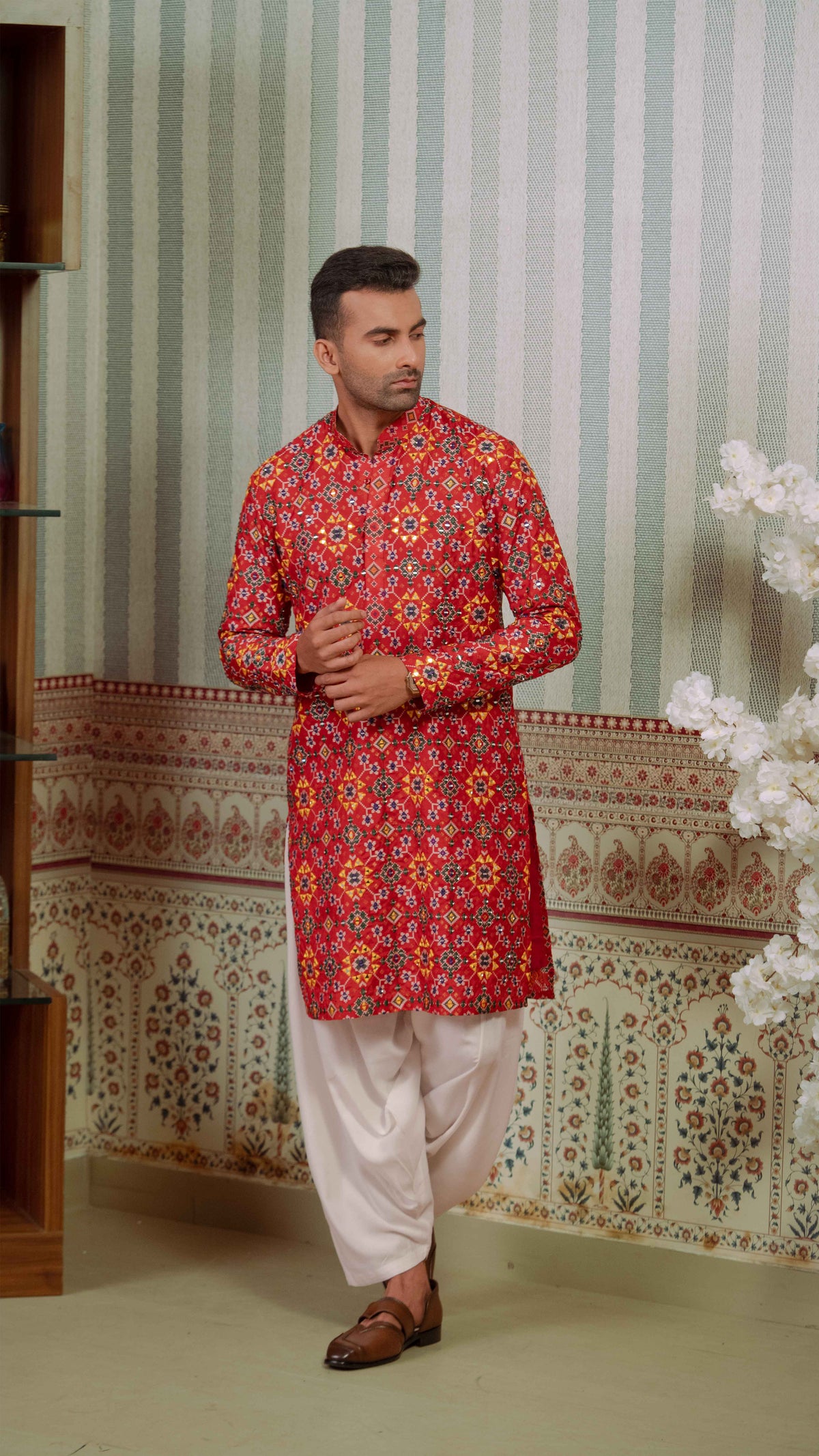 A man wearing a red and orange patterned kurta with white pants and brown sandals, standing in front of a patterned wallpaper background.