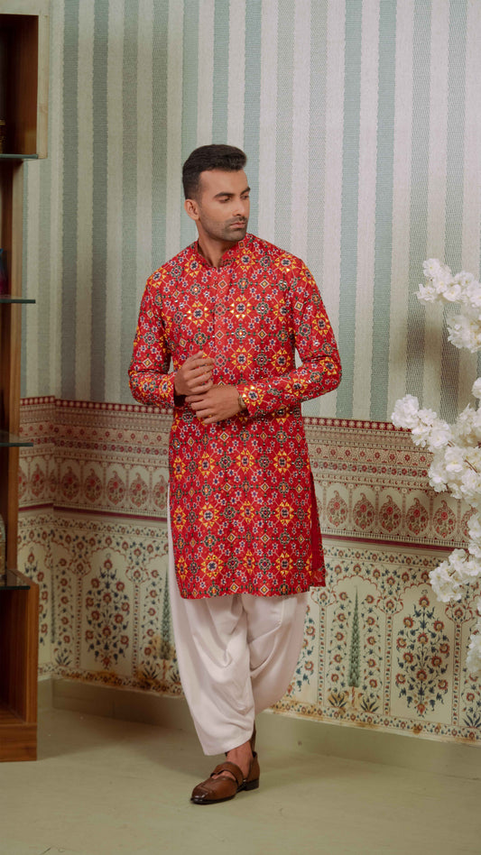 A man wearing a red and orange patterned kurta with white pants and brown sandals, standing in front of a patterned wallpaper background.