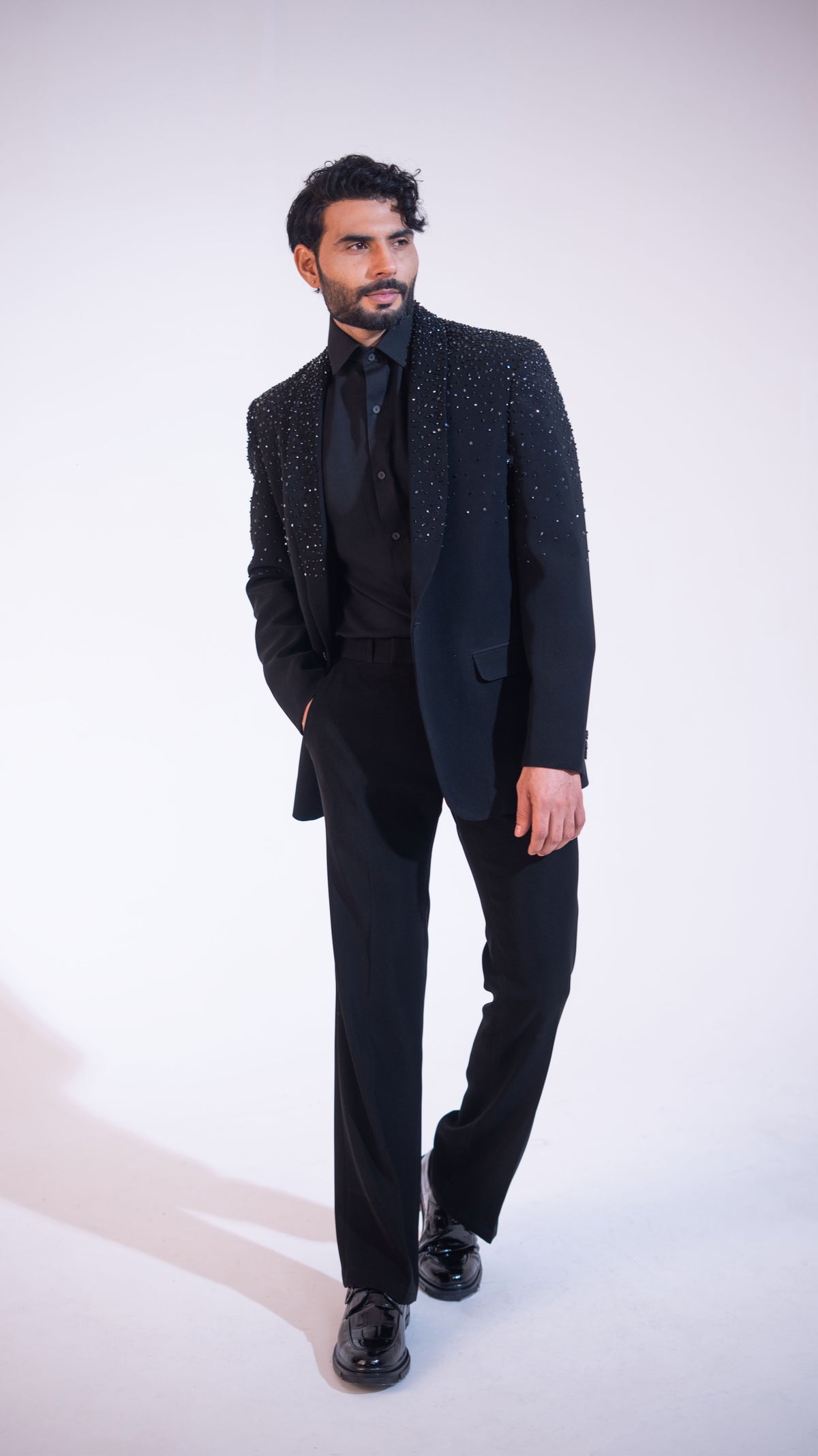 A man modeling a black formal suit with a heavily embellished, beaded jacket, worn with a black shirt, tie, and trousers, standing against a plain white studio background.