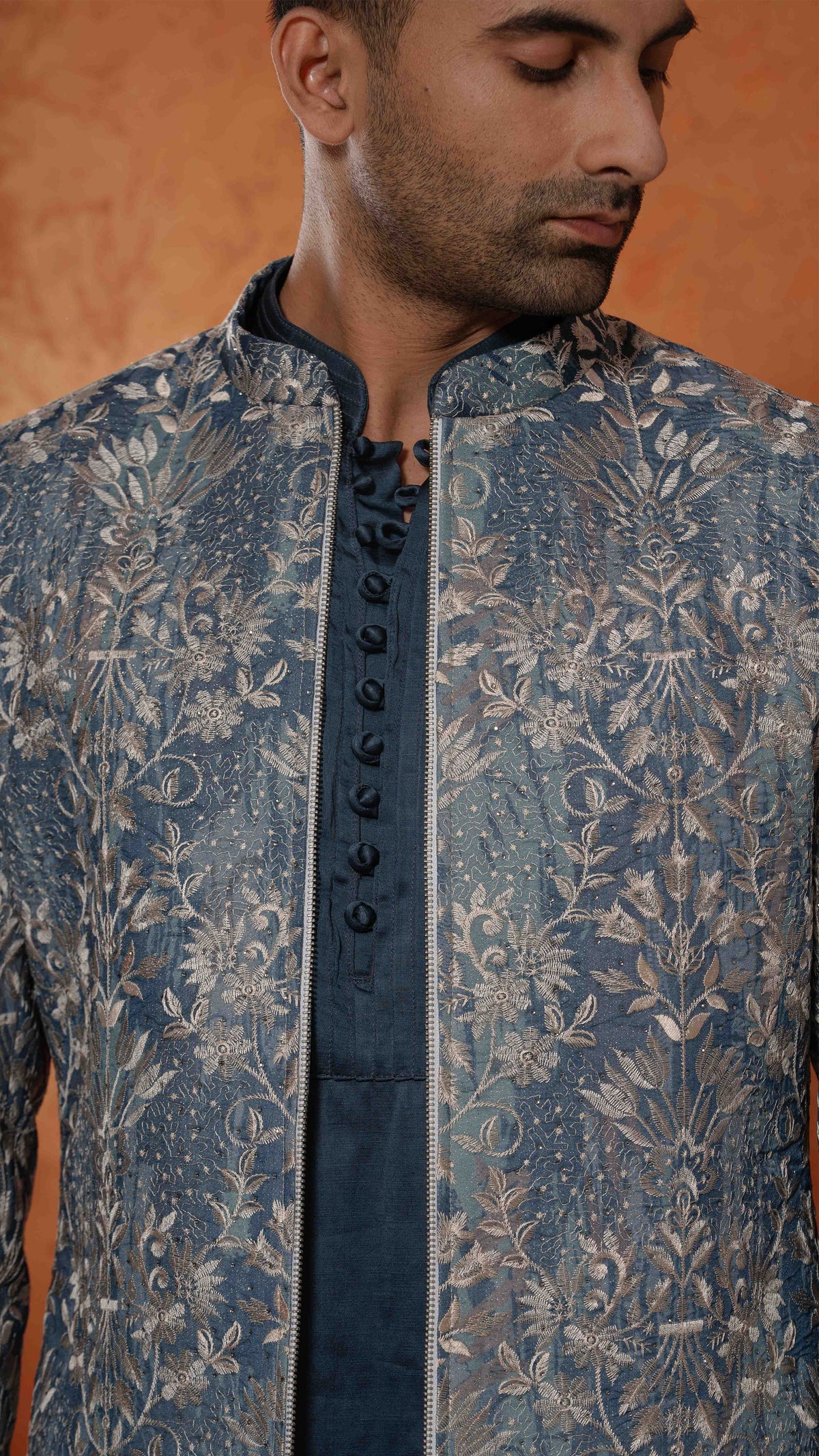 A close-up of a man wearing a blue and silver embroidered sherwani jacket with a matching dark blue inner shirt. The jacket features intricate floral embroidery and a central lace-up front panel.