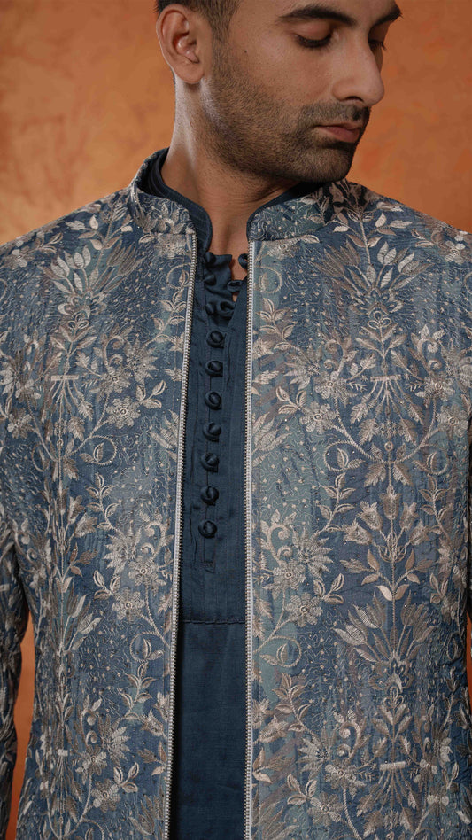 A close-up of a man wearing a blue and silver embroidered sherwani jacket with a matching dark blue inner shirt. The jacket features intricate floral embroidery and a central lace-up front panel.