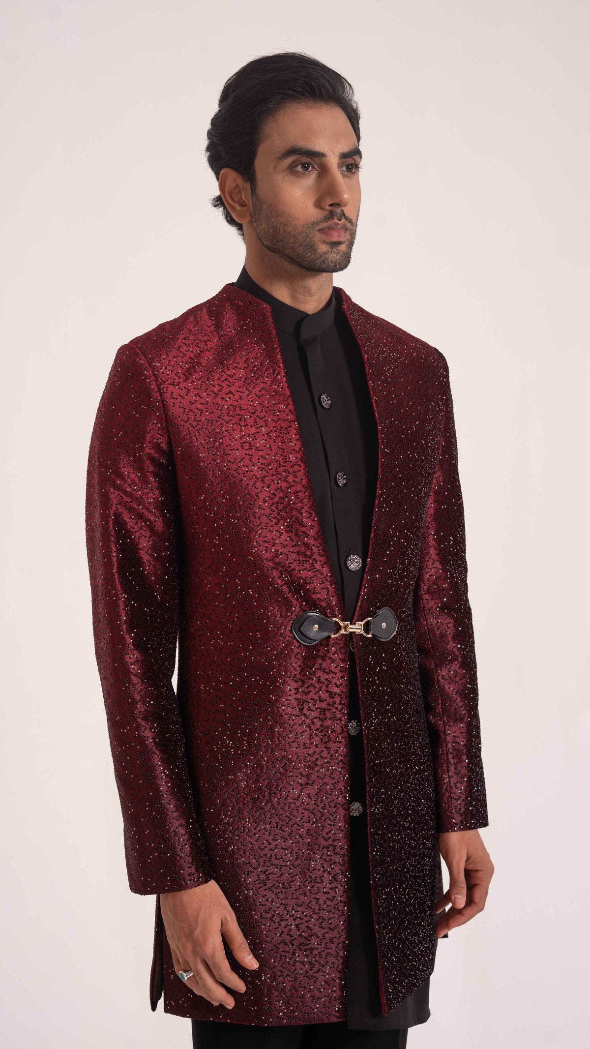 A man wearing a long-sleeved, open-front sherwani jacket in a deep maroon brocade pattern over a black kurta, standing against a plain light background.