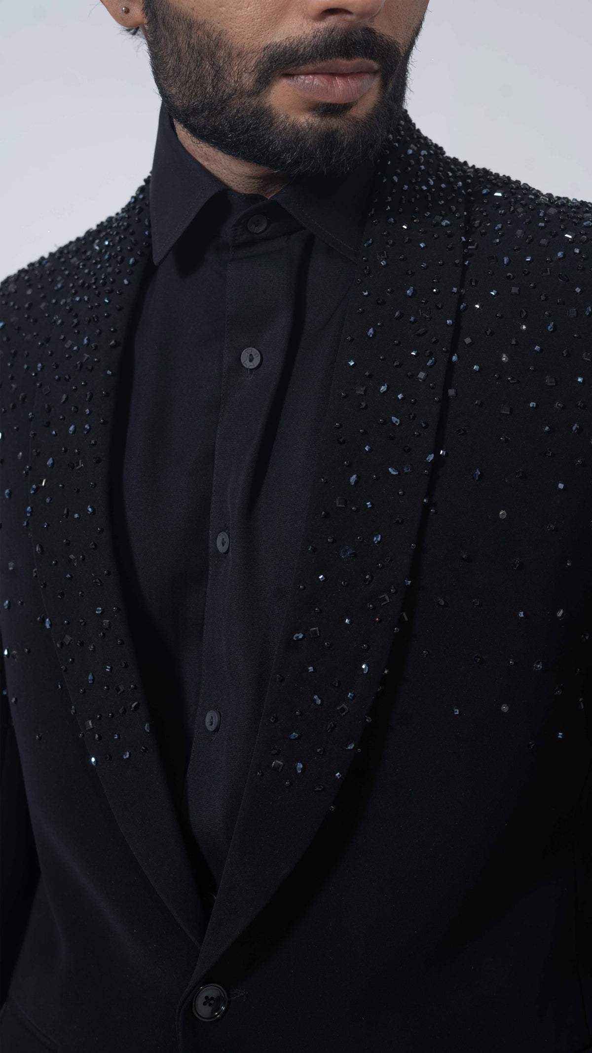 Close-up of a man wearing a black tailored jacket heavily embellished with sparkling silver and blue rhinestones over a black collared shirt, against a light gray studio background.
