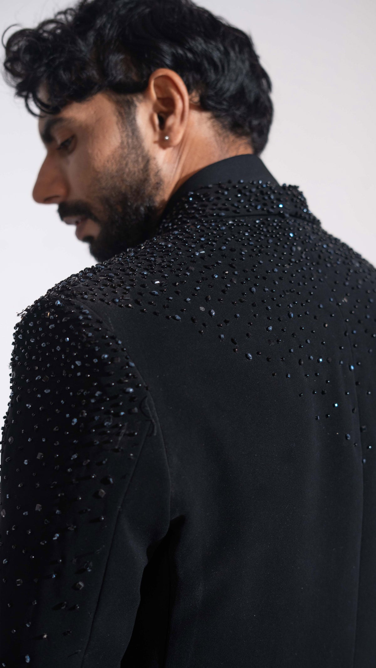 A man shown from the back and side wearing a black formal jacket heavily embellished with sparkling dark blue and silver sequins and beads on the shoulders and upper back, against a plain light gray studio background.