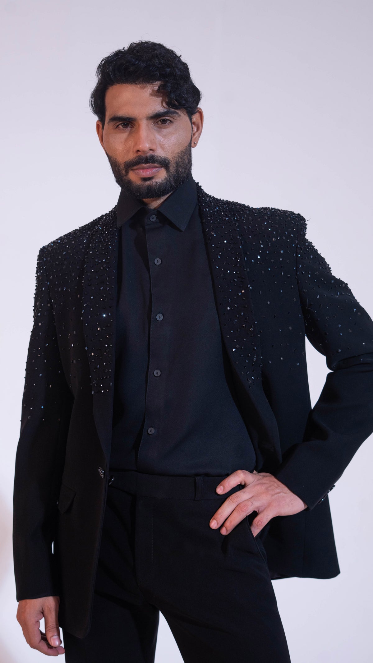 A man wearing a black embellished blazer over a black shirt and trousers, posing against a light gray studio background.