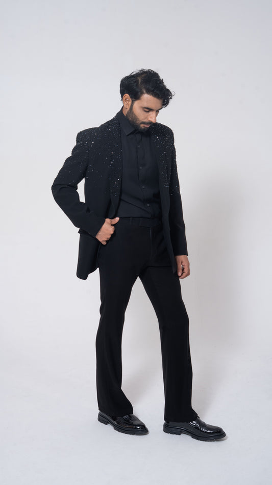 A man modeling a black formal suit with sparkling embellished lapels, a black shirt, black trousers, and black dress shoes against a light gray studio background.