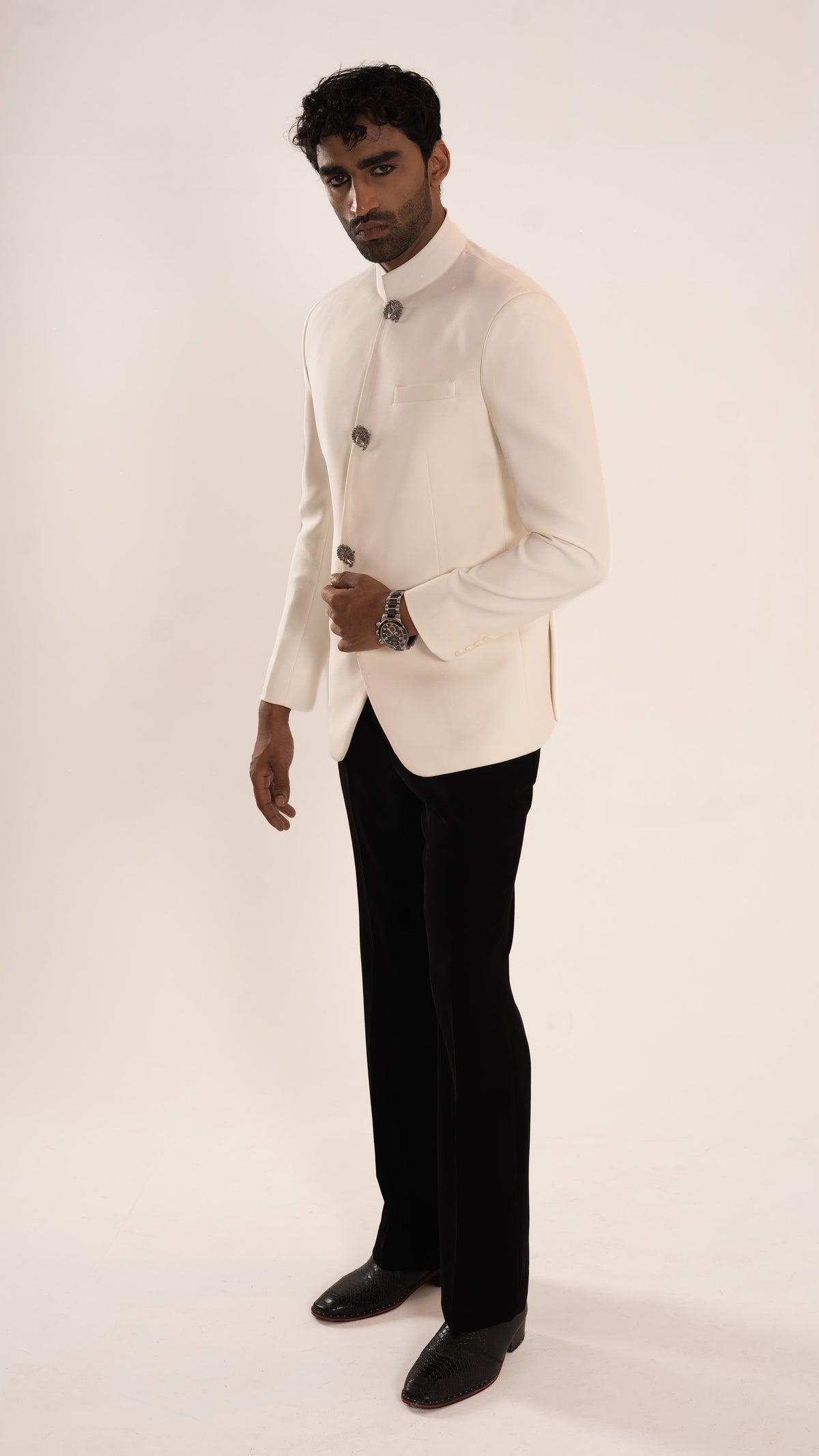 A man modeling a traditional white Indian jacket (jodhpuri) with black trousers and black shoes, standing against a plain off-white studio background.